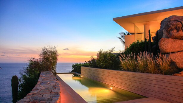 Una piscina moderna con bordo in pietra, un pontile in legno e vista sul mare al tramonto.