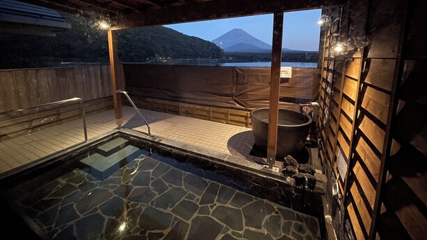 A hot spring bath area with a stone floor, wooden benches, and a view of a mountain. - Shoji Lake Hotel (Fujikawaguchiko)