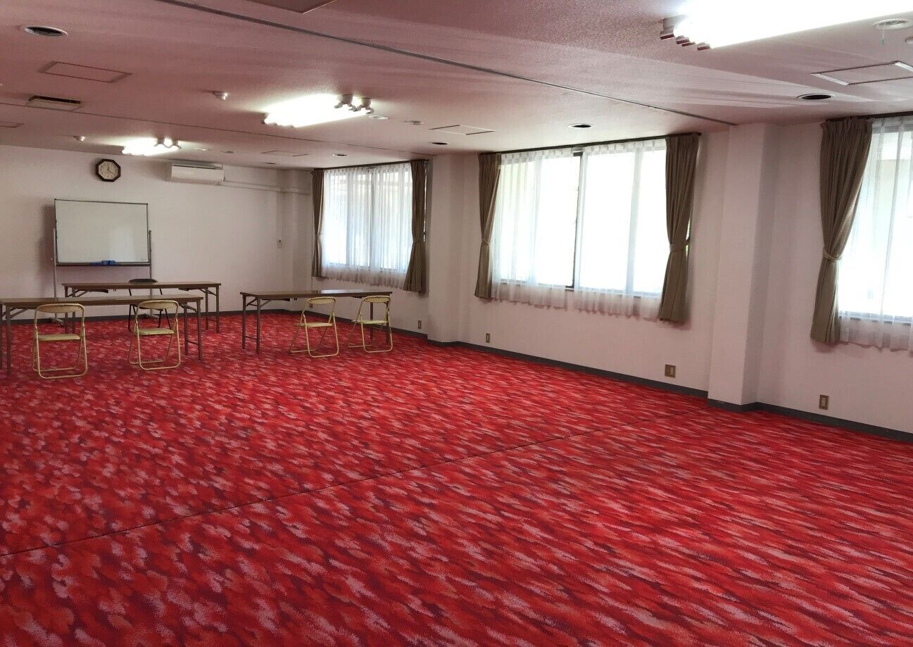 Un salón amplio con alfombra roja, una pizarra y varias mesas y sillas.