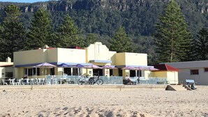 Een gebouw aan het strand met terras en parasols, omgeven door bomen en bergen.
