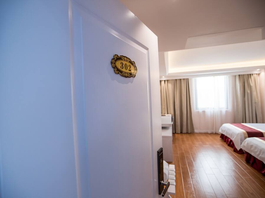 A hotel room with a wooden floor, two beds with red and white bedding, a window with beige curtains, and a wall-mounted number plate.