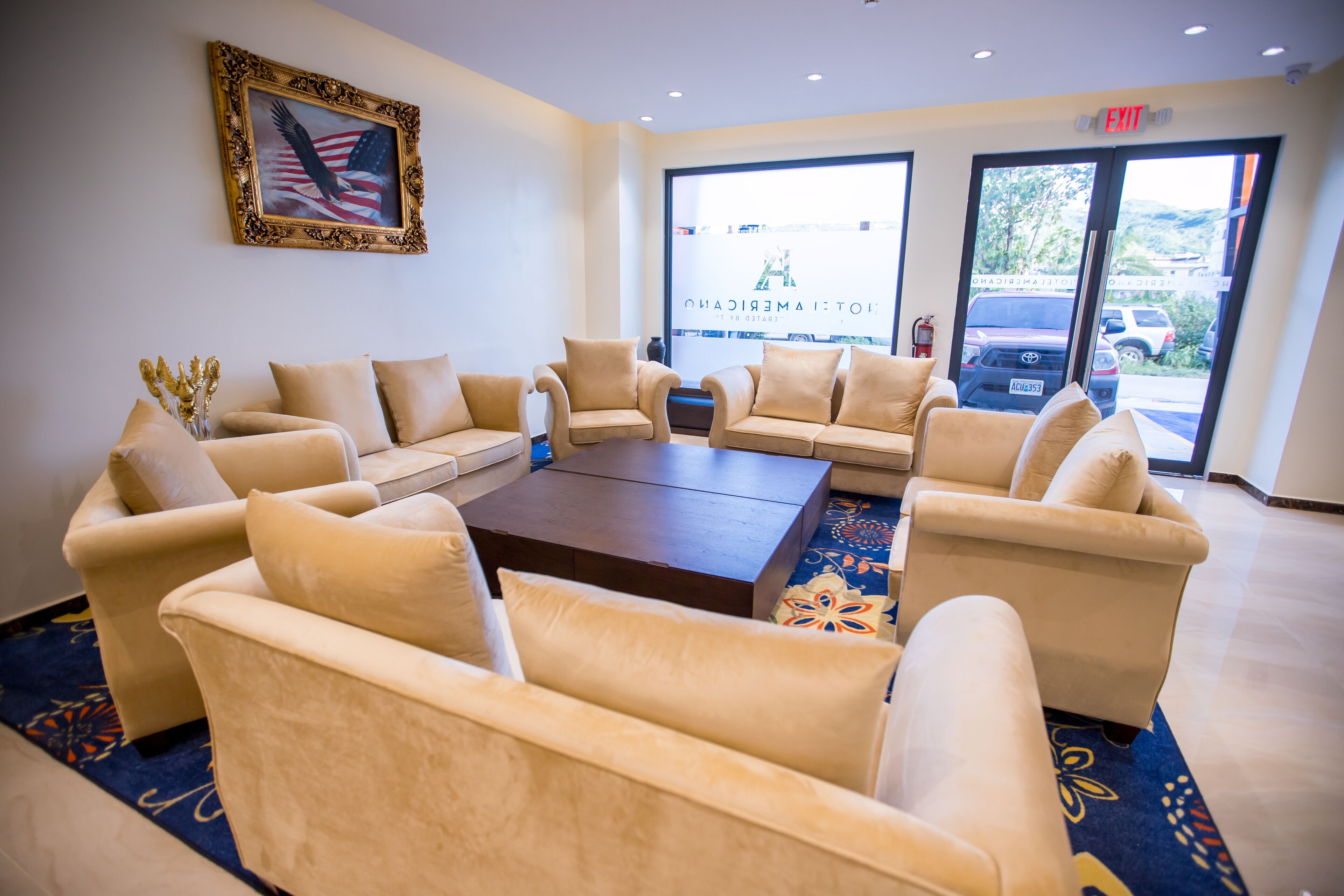 Een hal met beige banken rond een centrale salontafel, een Amerikaanse vlag in een lijst aan de muur, en een uitzicht op geparkeerde auto's door grote ramen.