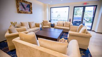 Een hal met beige banken rond een centrale salontafel, een Amerikaanse vlag in een lijst aan de muur, en een uitzicht op geparkeerde auto's door grote ramen.