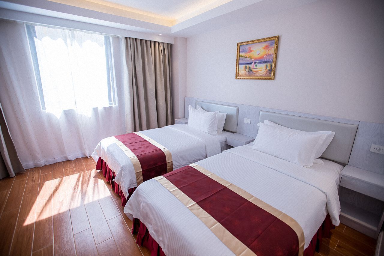 A hotel room with two beds, a painting on the wall, and a large window with curtains.