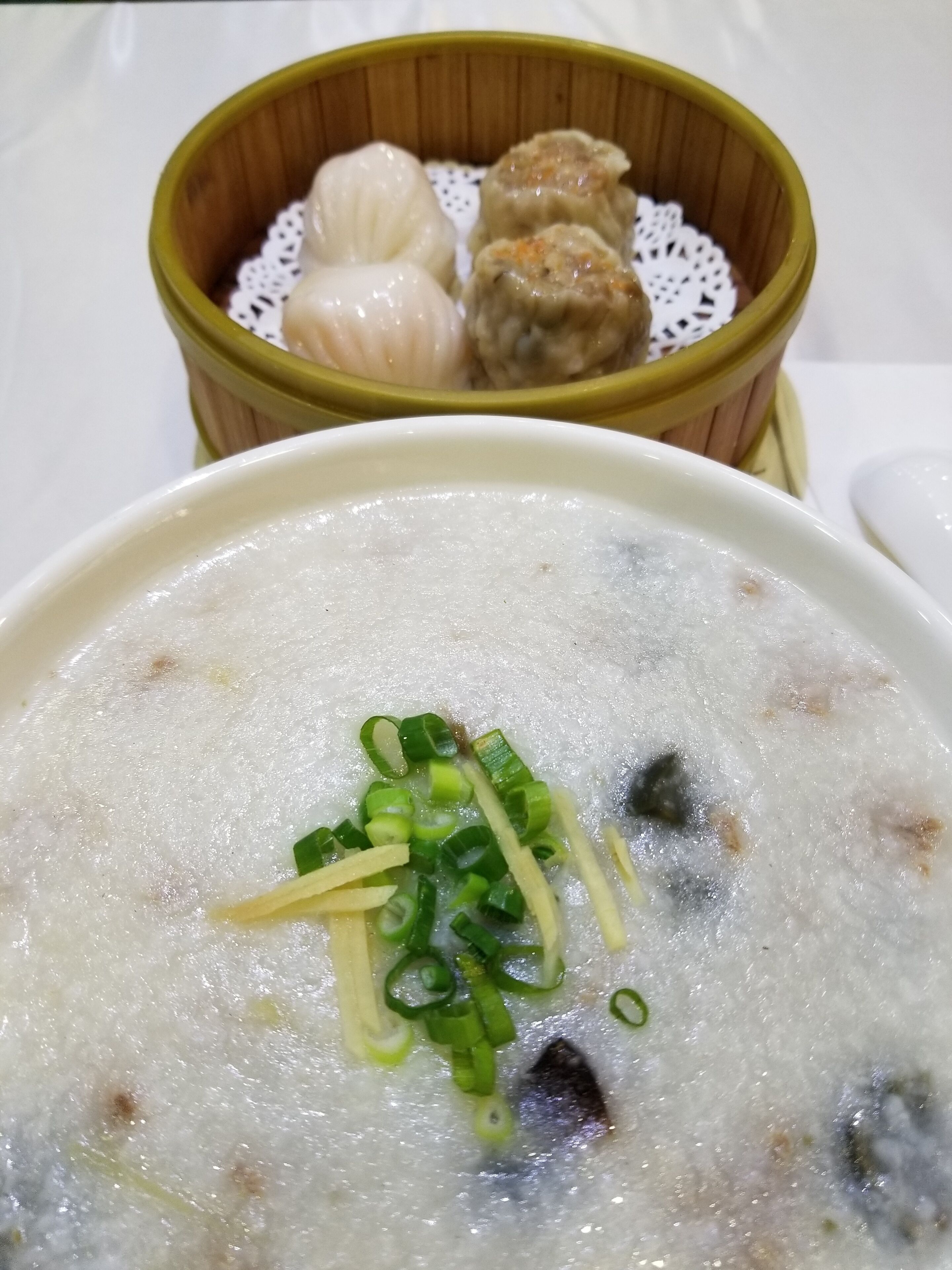 Un tazón de gachas de arroz con cebolletas y una vaporera de bambú con dumplings al fondo.