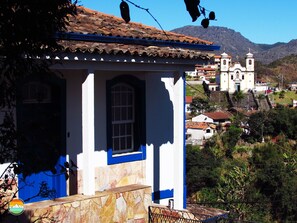 Un edificio blanco con detalles azules y base de piedra, una torre de campanas y montañas al fondo.