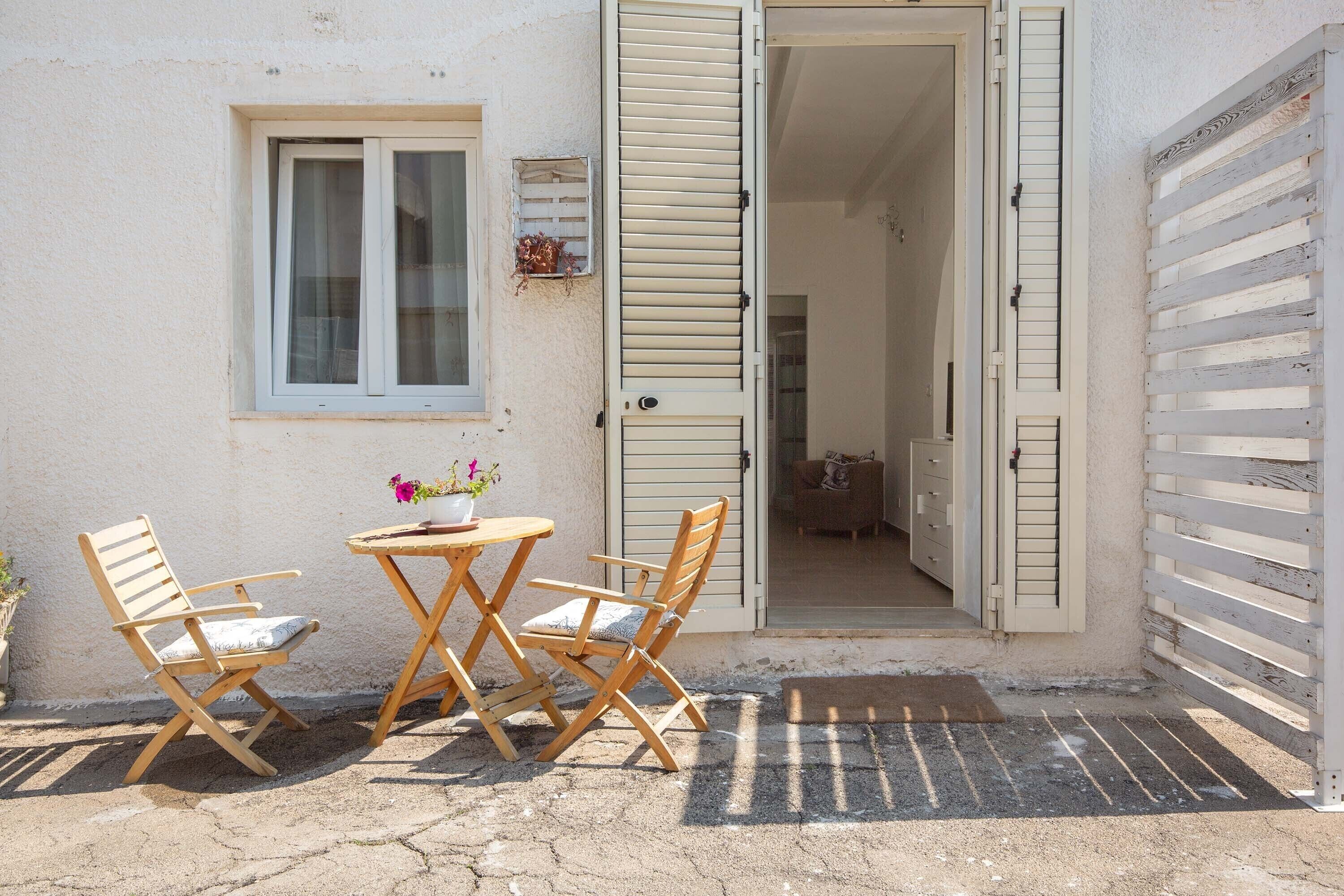 An outdoor seating area with two wooden chairs and a table, a window with shutters, and a door leading inside.