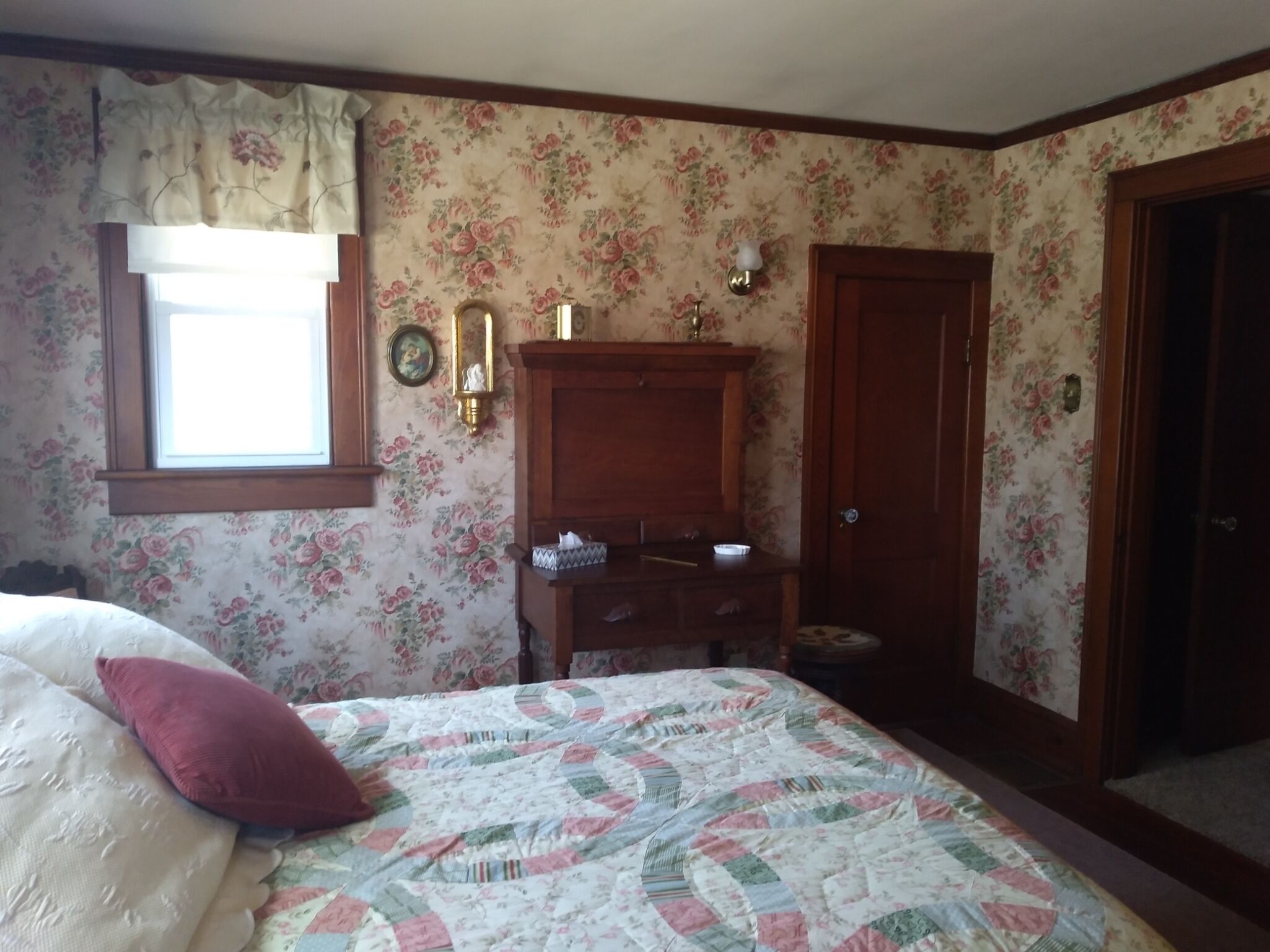 A bedroom with floral wallpaper, a wooden dresser, a bed with a quilt, and a window with a curtain.