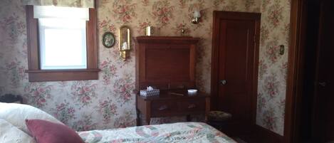 Un dormitorio con papel tapiz floral, un aparador de madera, una cama con colcha y una ventana con cortina.