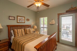 A bedroom with a wooden bed, a nightstand, a lamp, a ceiling fan, a window with blinds, and a door with a small sailboat decoration.