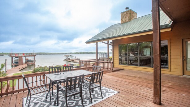 Um deck de madeira com mesa e cadeiras, com vista para um corpo d'água com barcos e um cais.