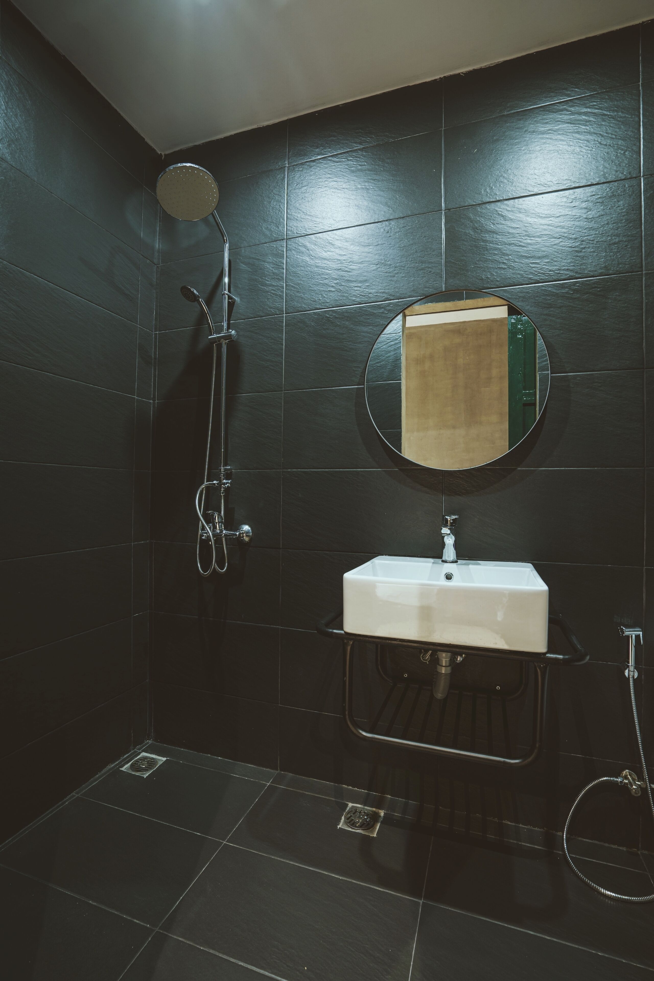 Une salle de bain avec des murs recouverts de carreaux foncés, un miroir rond, un lavabo blanc et une douche équipée d’un pommeau de douche à main.