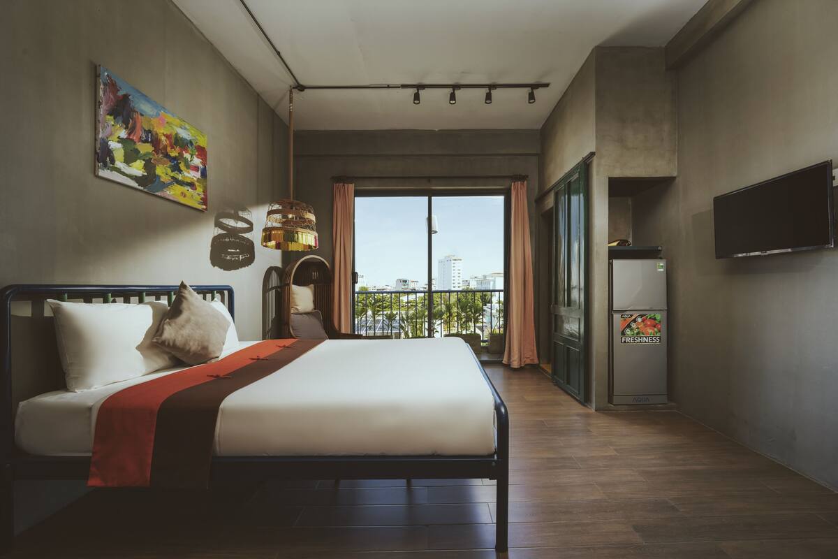 A modern hotel room with a large bed, a TV, a balcony with a city view, and a refrigerator.