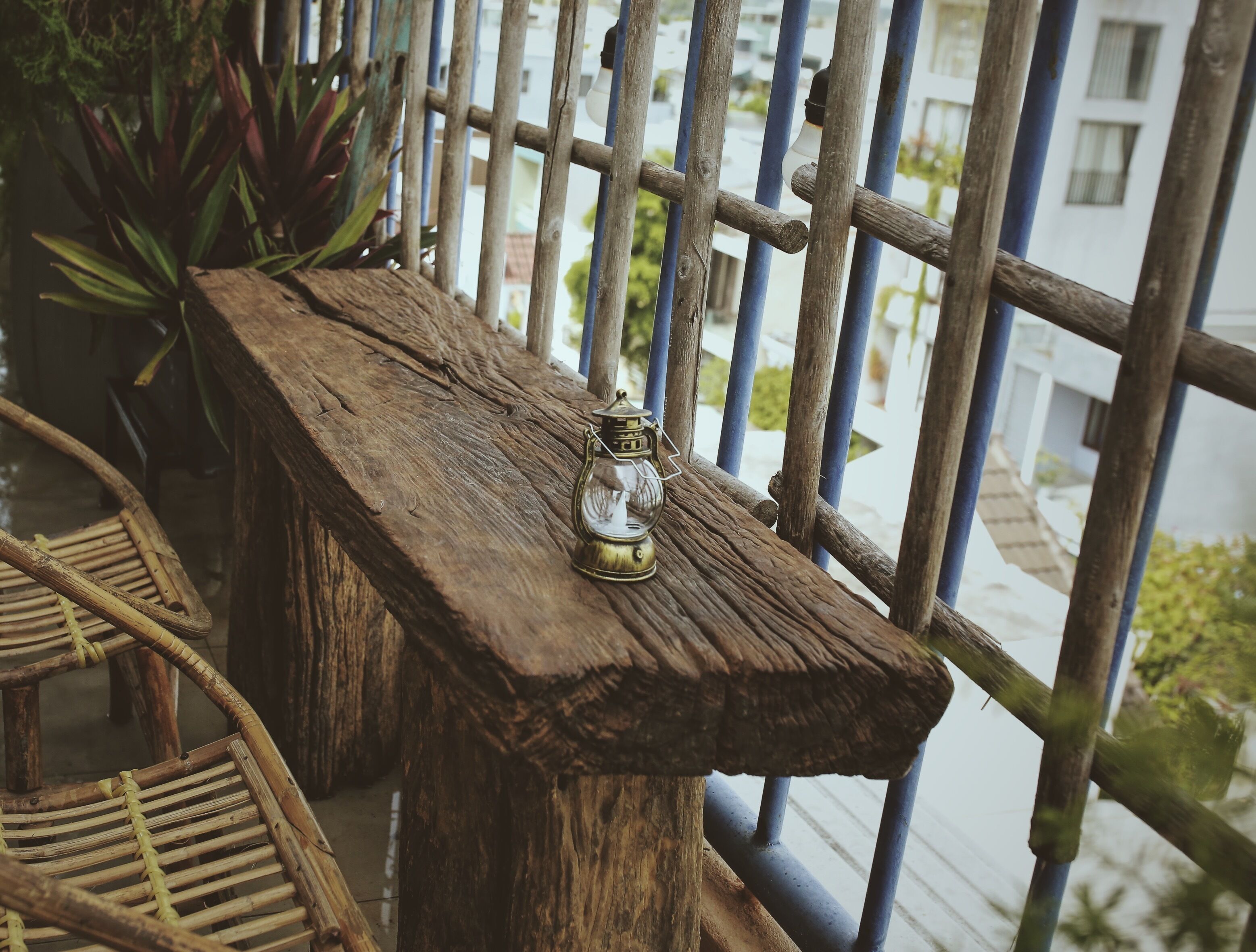A rustic wooden bench with a lantern on it, set against a metal railing with a view of buildings in the background.