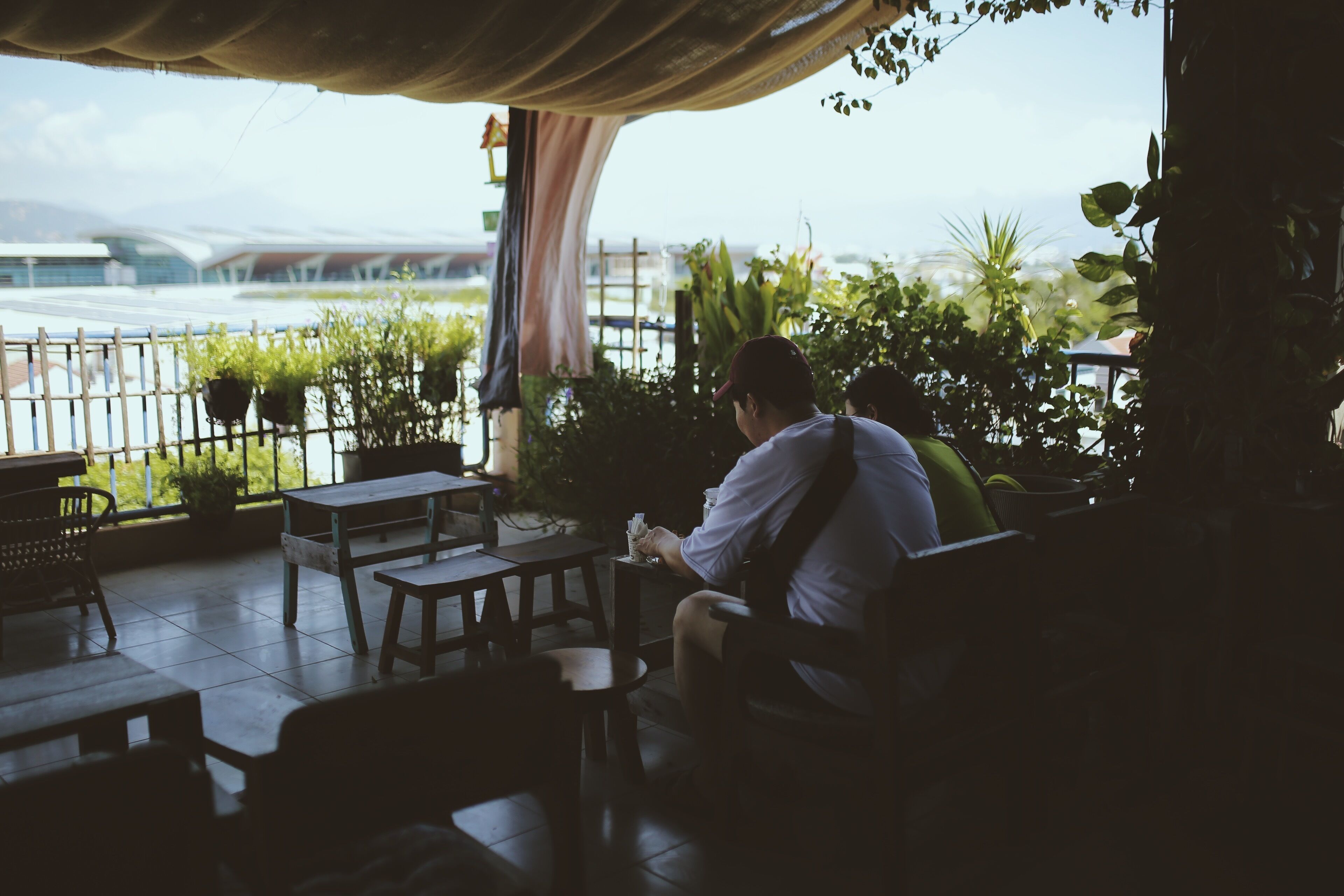 A person seated at a table on a covered patio with a view of a modern building and greenery.