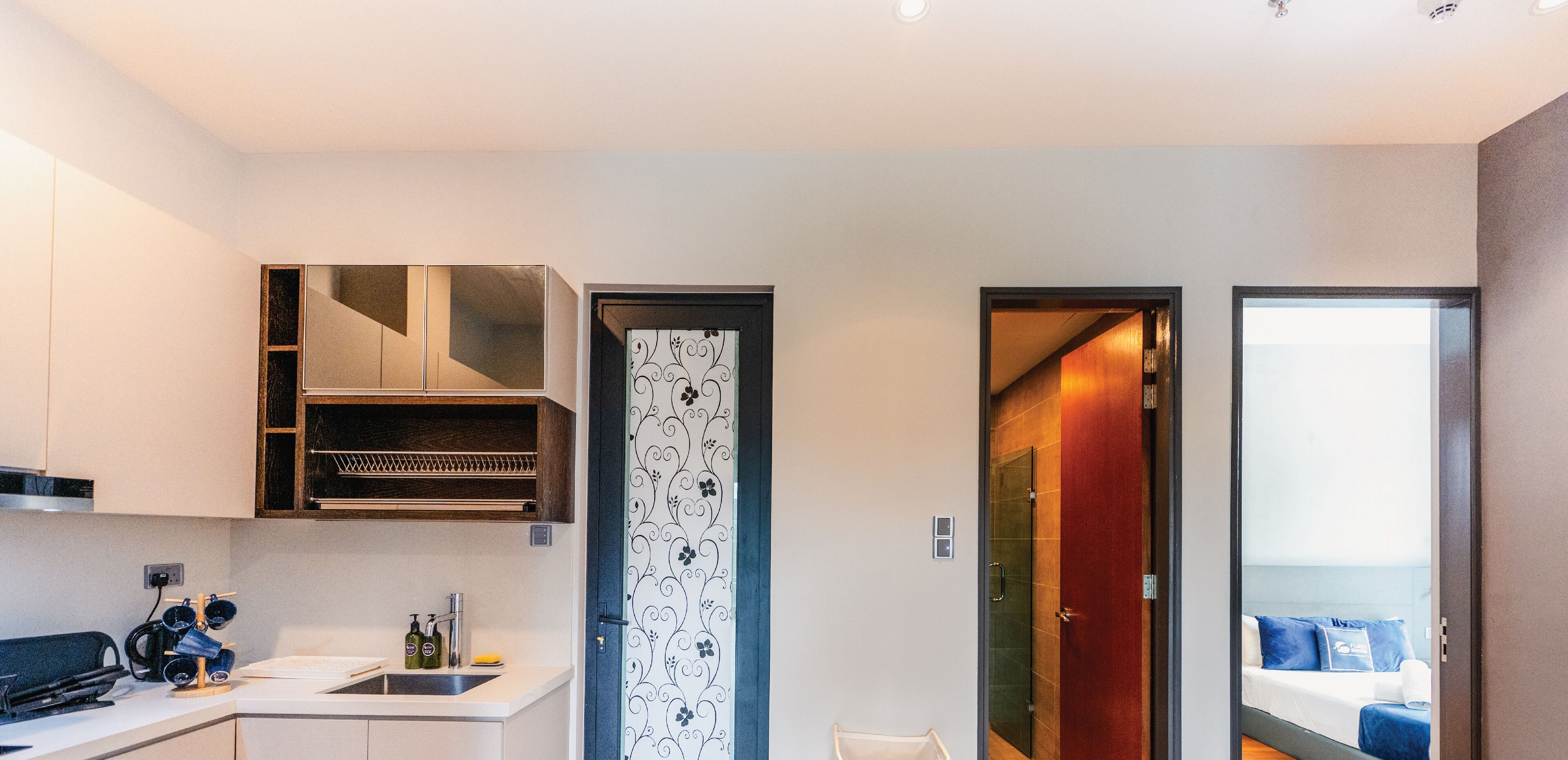 A modern kitchen with a built-in oven, a sink, and a countertop with a coffee machine and bottles. There is a door with a decorative pattern leading to a bedroom with a bed and a nightstand.