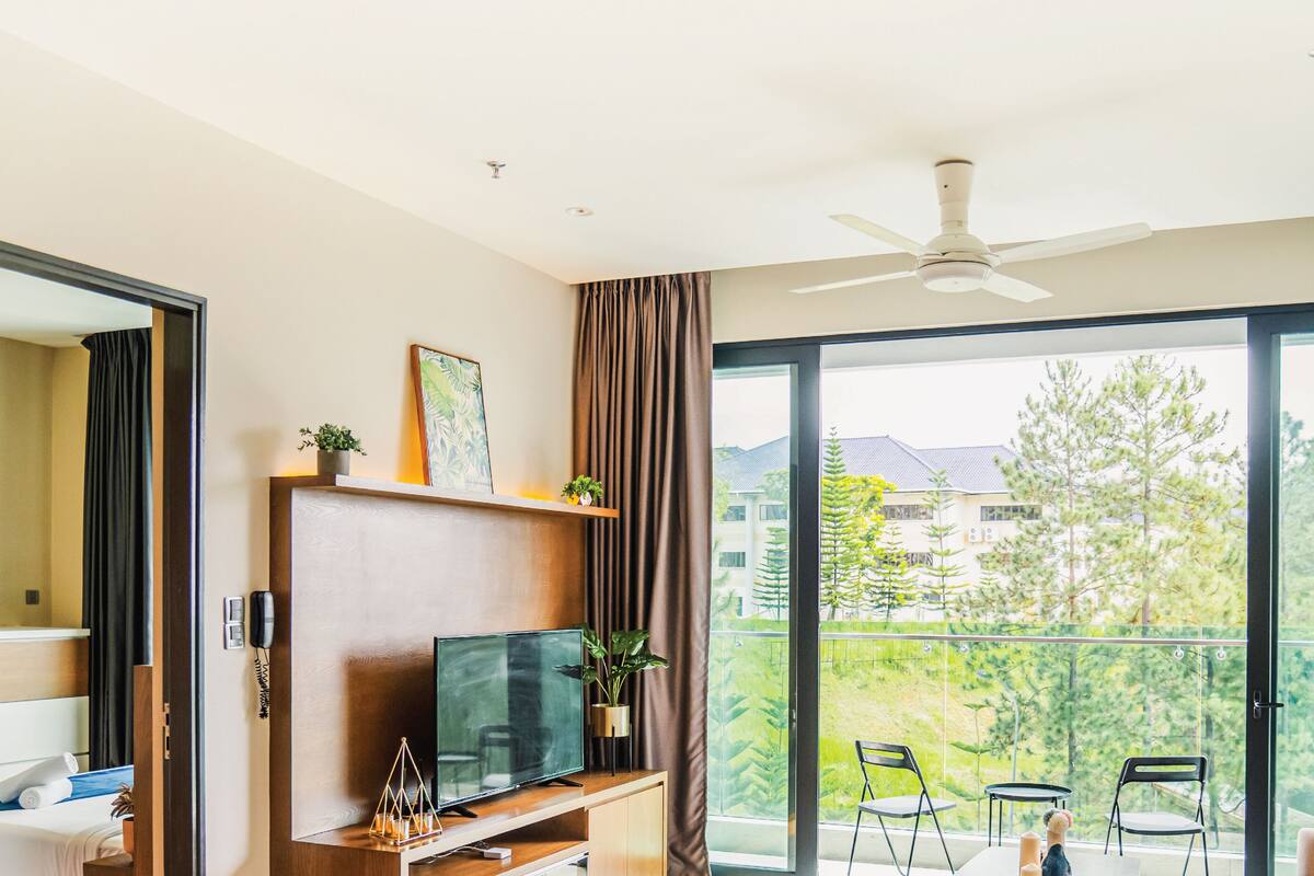 A modern hotel room with a large balcony, a flat-screen TV, a wooden entertainment unit, and a view of the surrounding area.