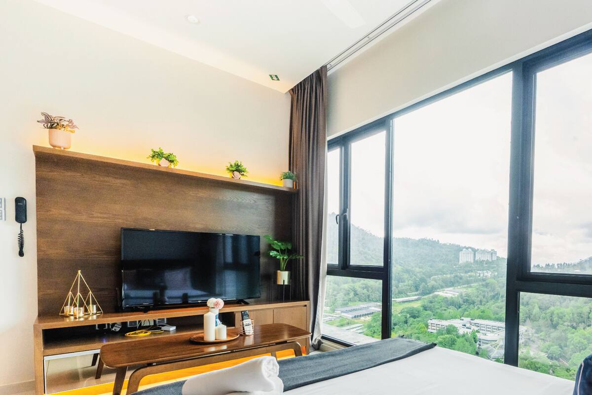 A hotel room with a large window, a flat-screen TV, a wooden desk, and a bed with pillows.