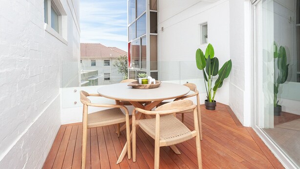 Uma varanda com uma mesa redonda de madeira e quatro cadeiras de vime, um vaso de plantas e uma porta de vidro que dá para um espaço interno.