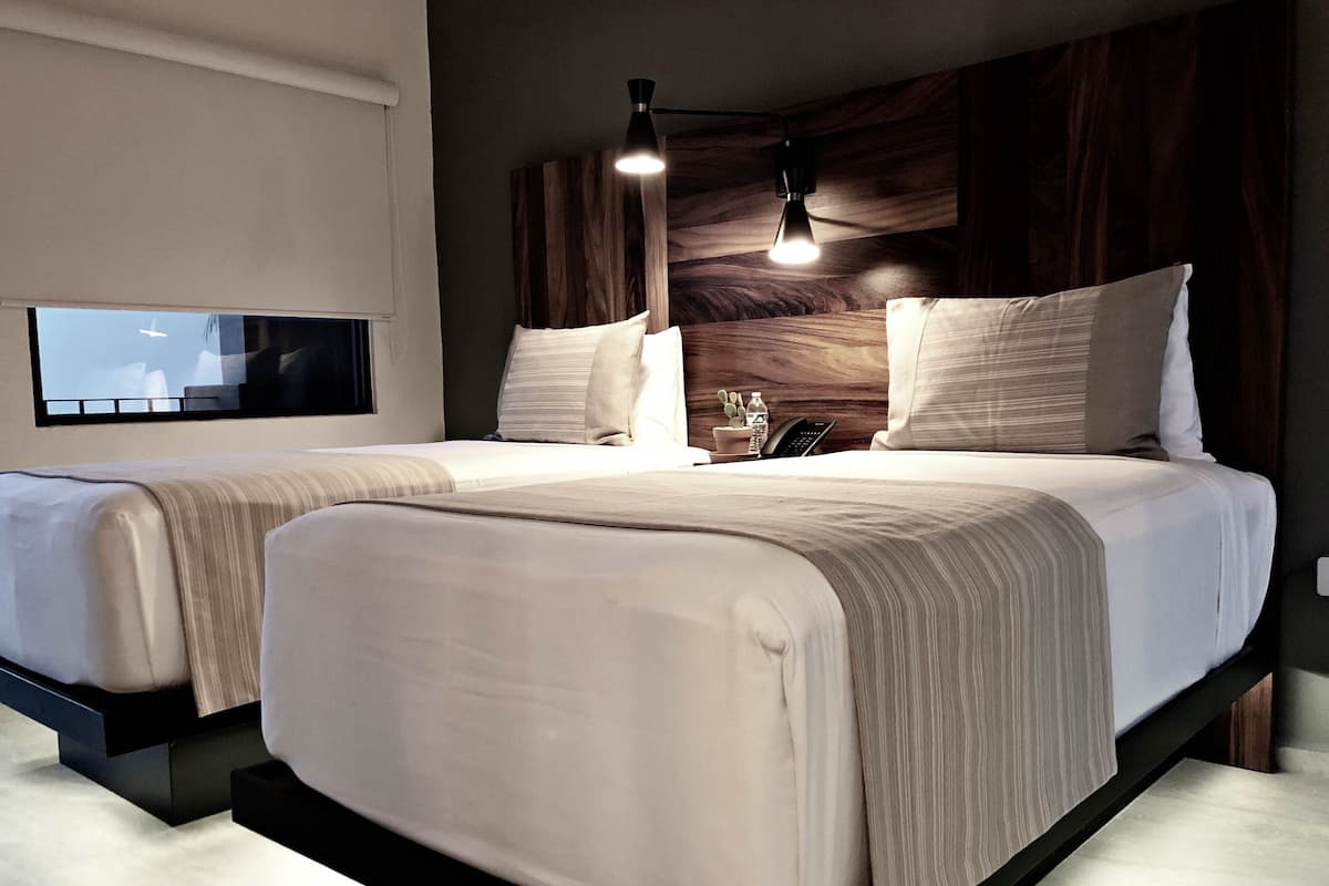 A modern hotel room with two beds, a flat-screen TV, a bedside table with a lamp, and a wooden headboard.