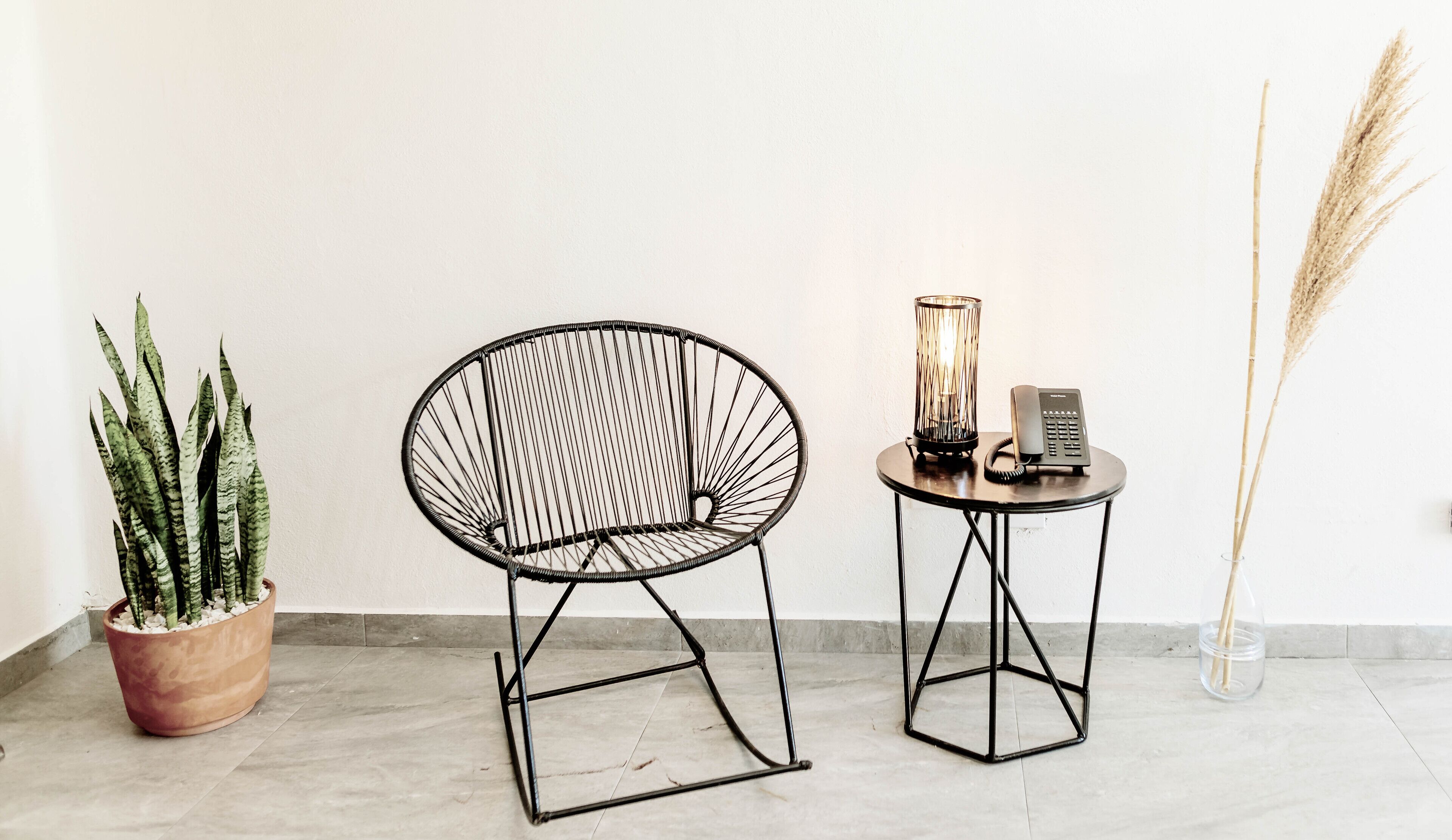 Una habitación con una silla de estructura metálica negra, una pequeña mesa redonda con un teléfono y una vela, y una planta en maceta con hojas altas y delgadas.
