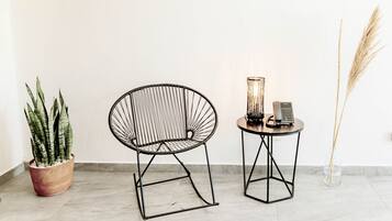 Una habitación con una silla de estructura metálica negra, una pequeña mesa redonda con un teléfono y una vela, y una planta en maceta con hojas altas y delgadas.