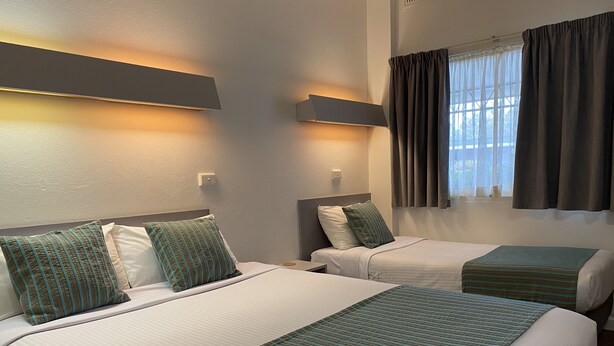 A hotel room with two beds, each with a striped bedspread and pillows. There are wall-mounted light fixtures above the beds and a window with curtains.