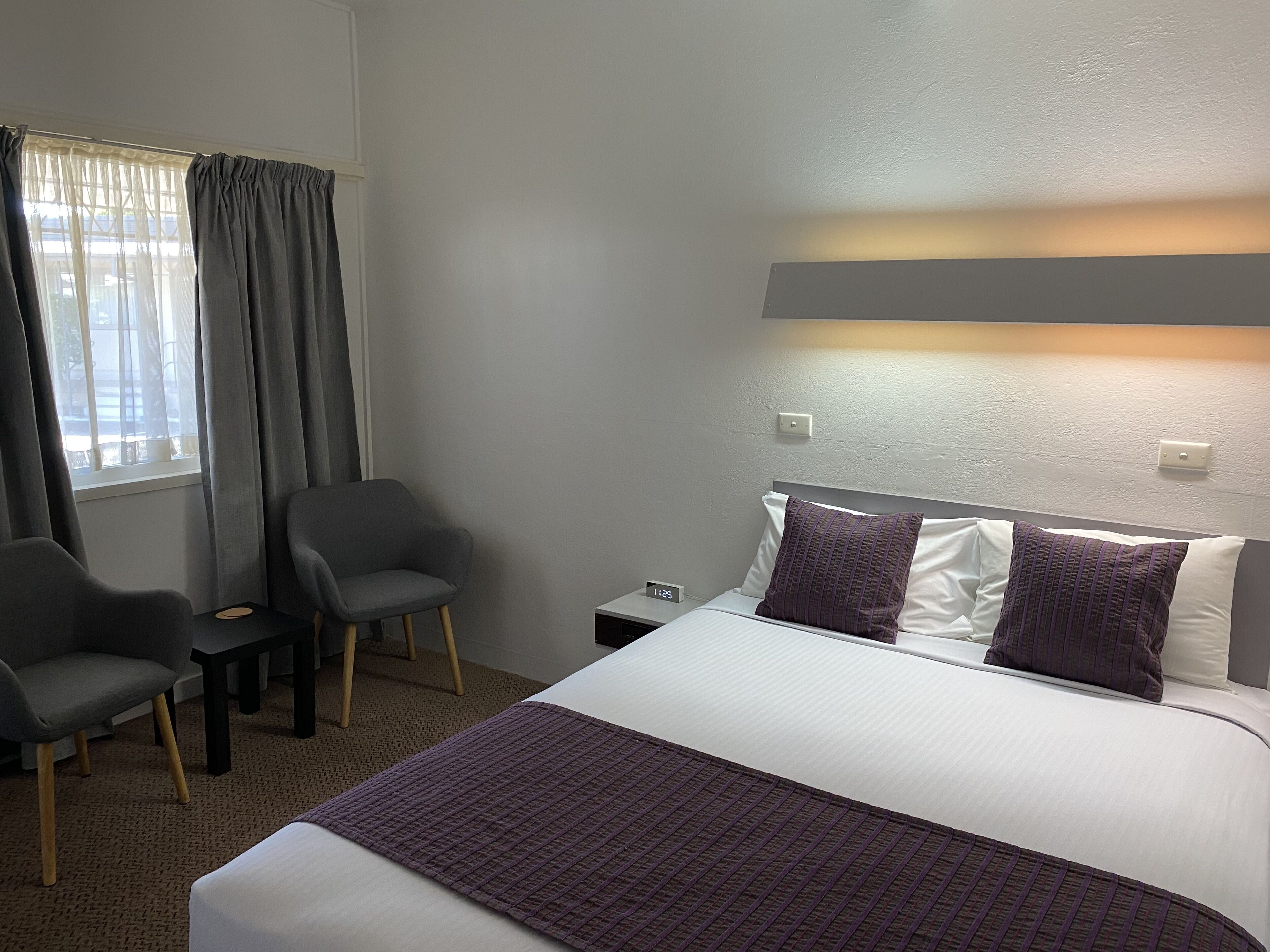A hotel room with a bed, a chair, a nightstand, and a window with curtains.