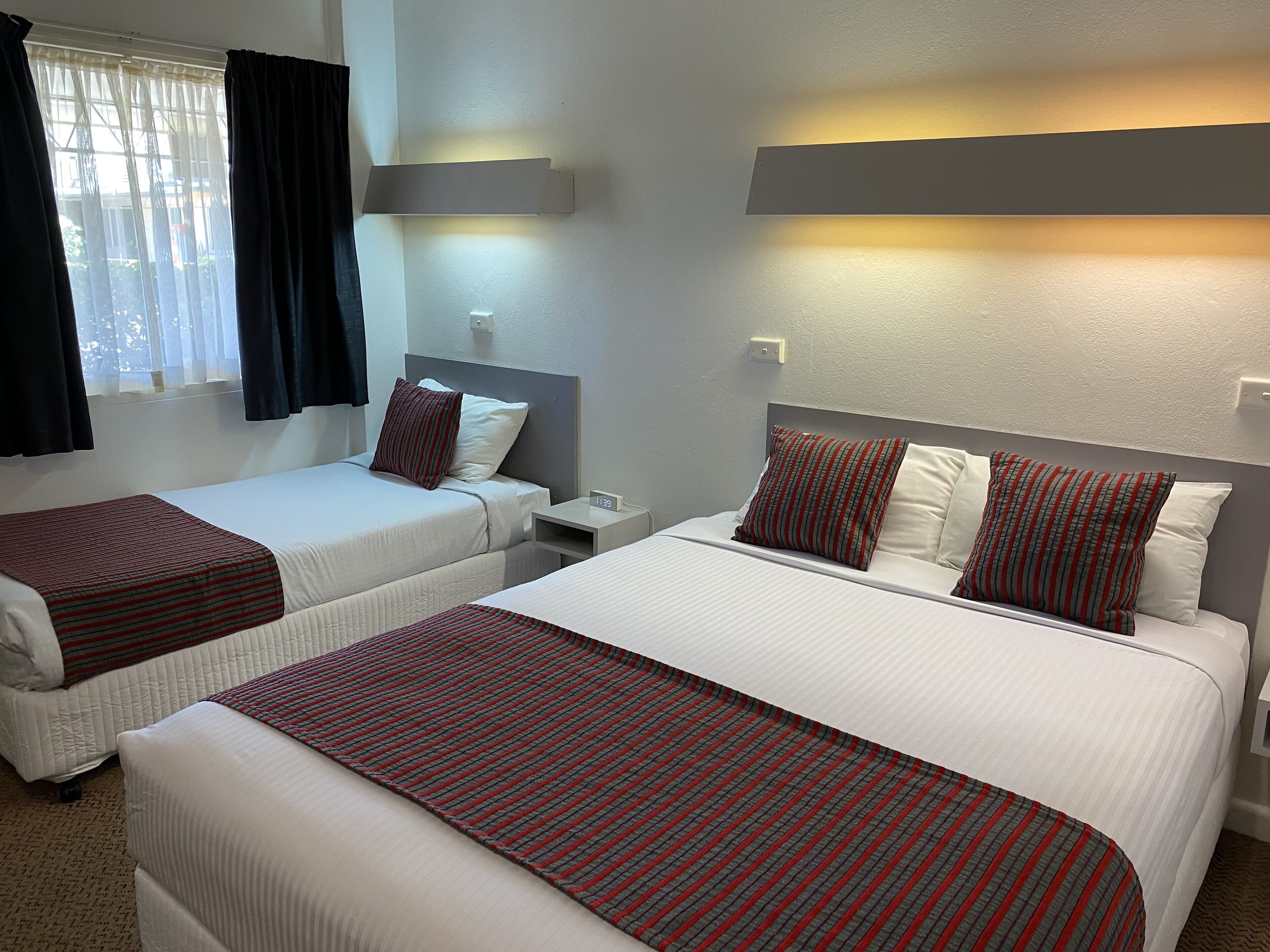 Une chambre d’hôtel avec deux lits, chacun recouvert d’une couvre-lit à rayures rouges et grises, agrémenté de draps blancs et d’oreillers assortis. Une table de chevet avec une lampe se trouve à côté de chaque lit, et un luminaire mural est fixé au-dessus des lits.