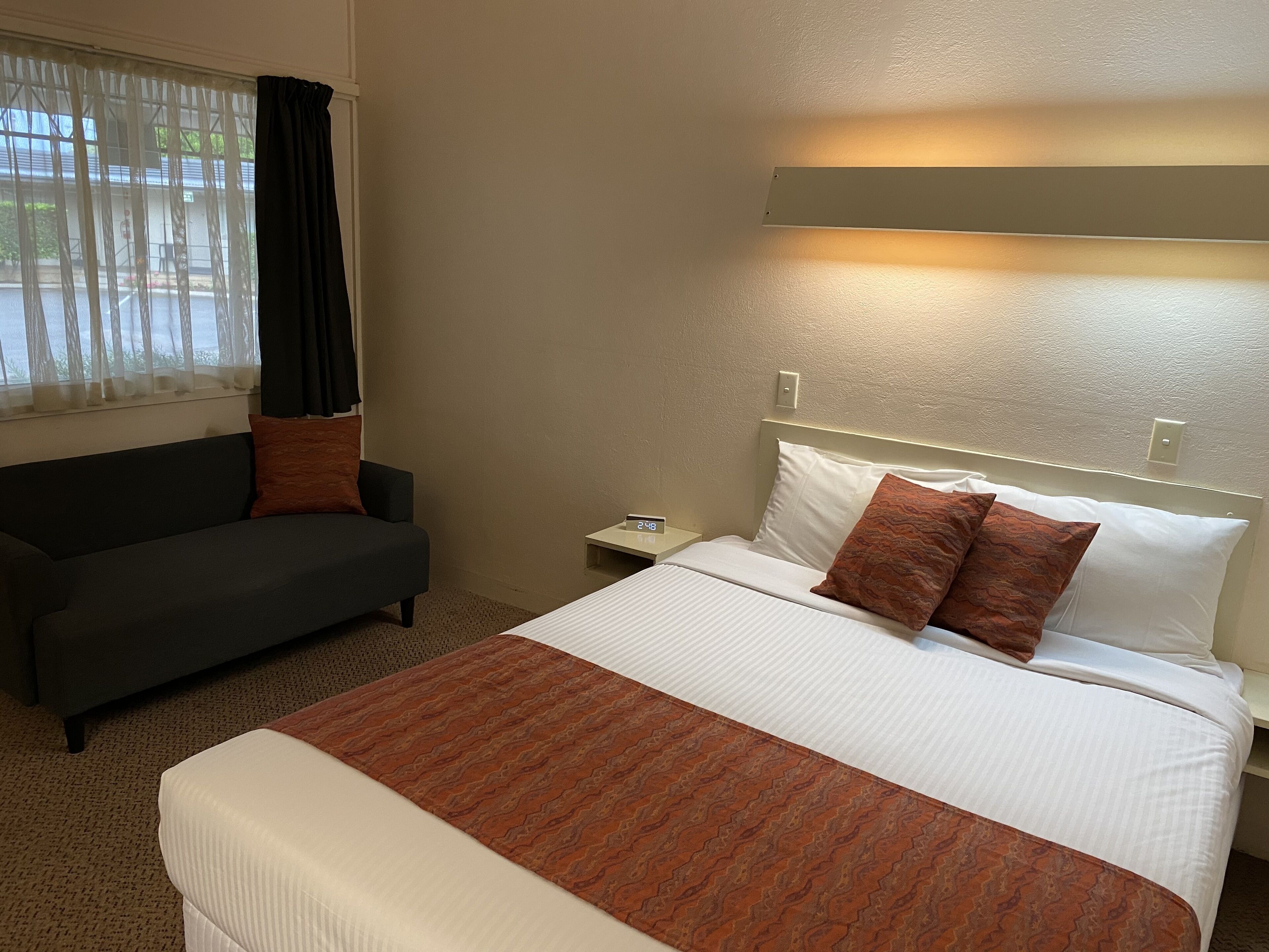 A hotel room with a bed, a nightstand, a sofa, and a window with curtains.
