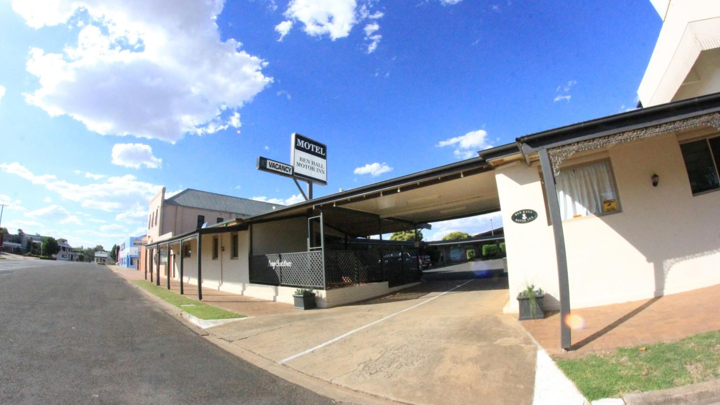 Um motel com área de estacionamento coberta, placa indicando quartos disponíveis e um edifício visível com uma placa de identificação.