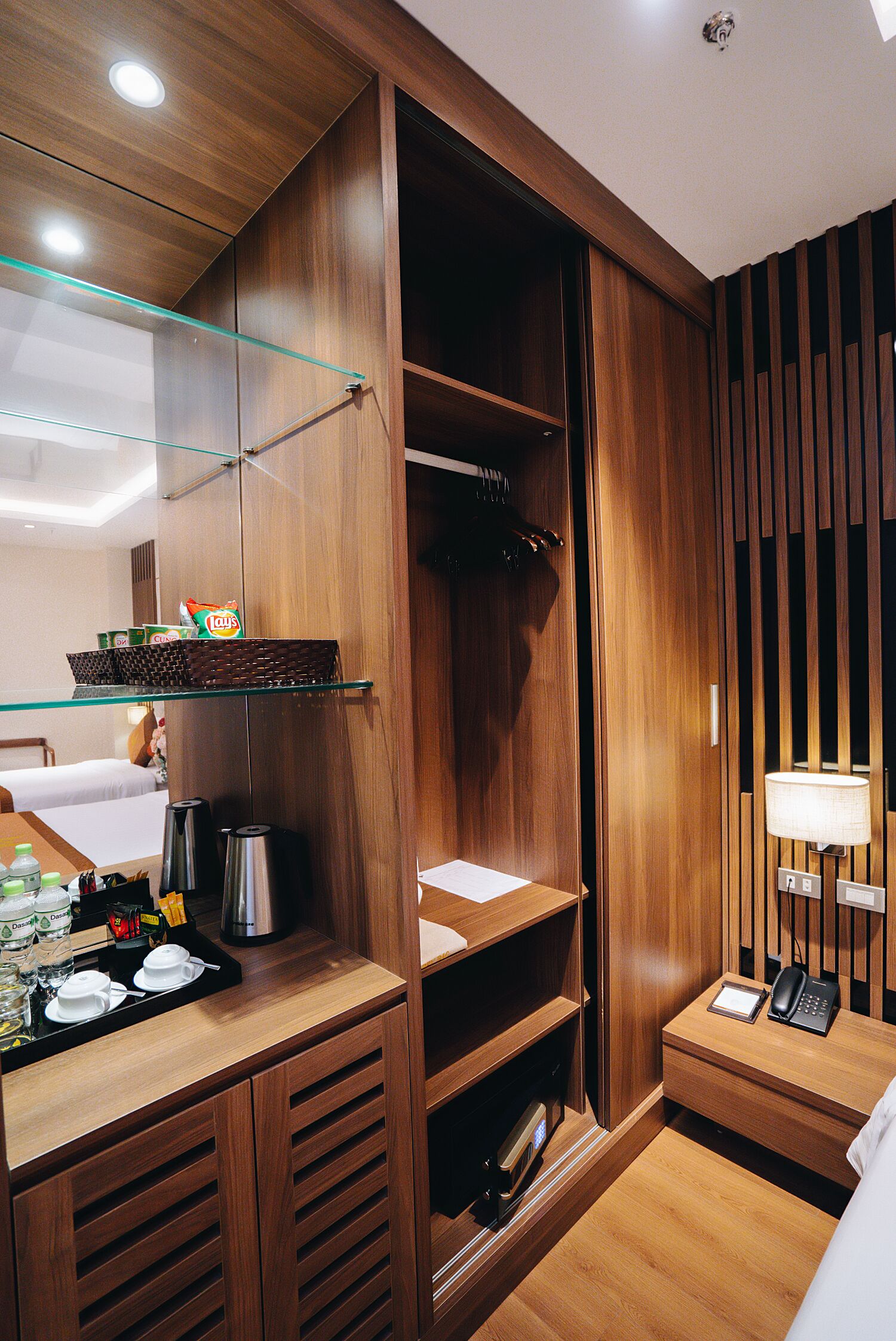 A hotel room with a wooden wardrobe, a bedside table with a lamp, and a small shelf with toiletries.