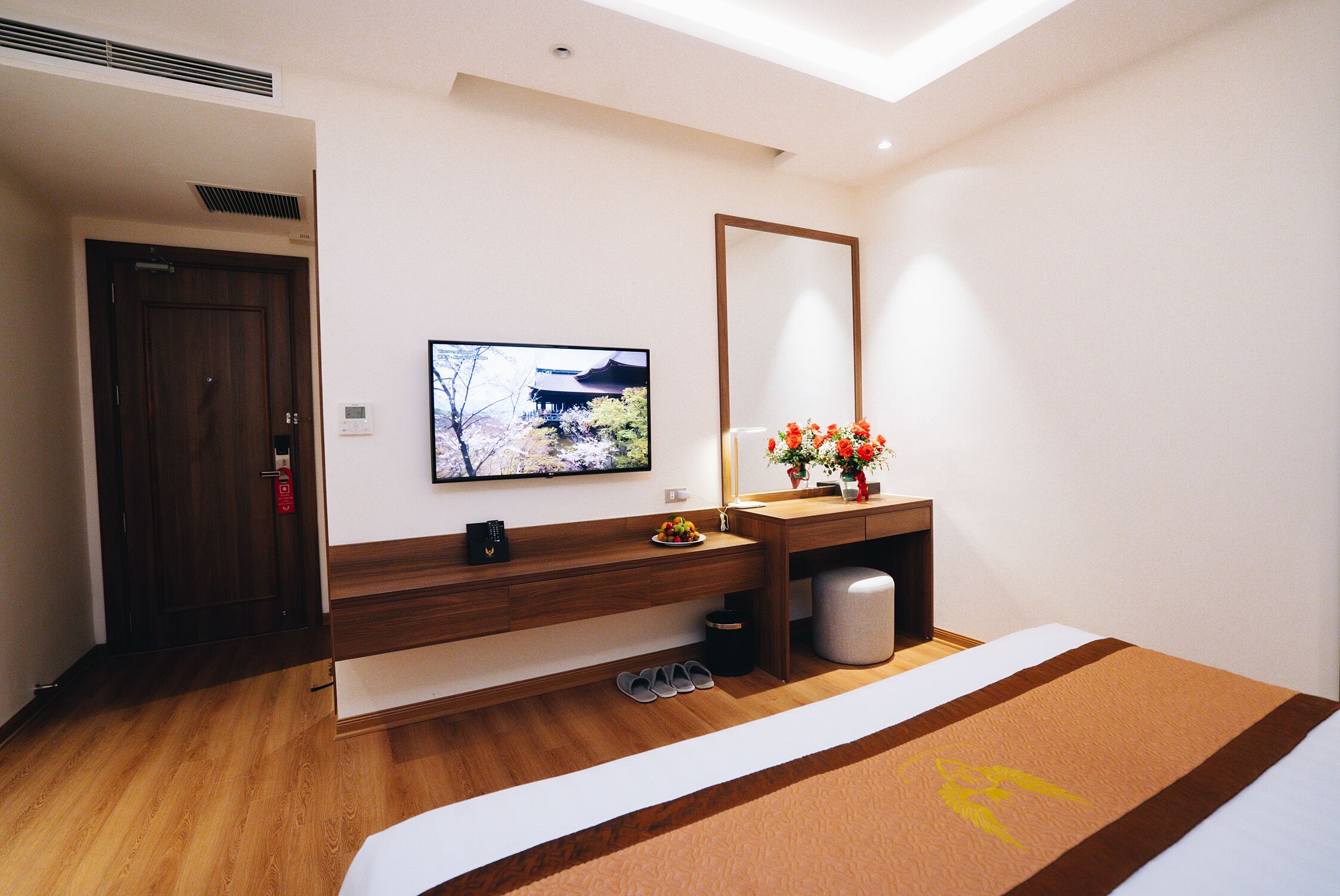 Una camera d'albergo con pavimento in legno, un letto con copripiumino marrone e bianco, una televisione fissata al muro, una scrivania in legno con un vaso di fiori e uno specchio sopra la scrivania.