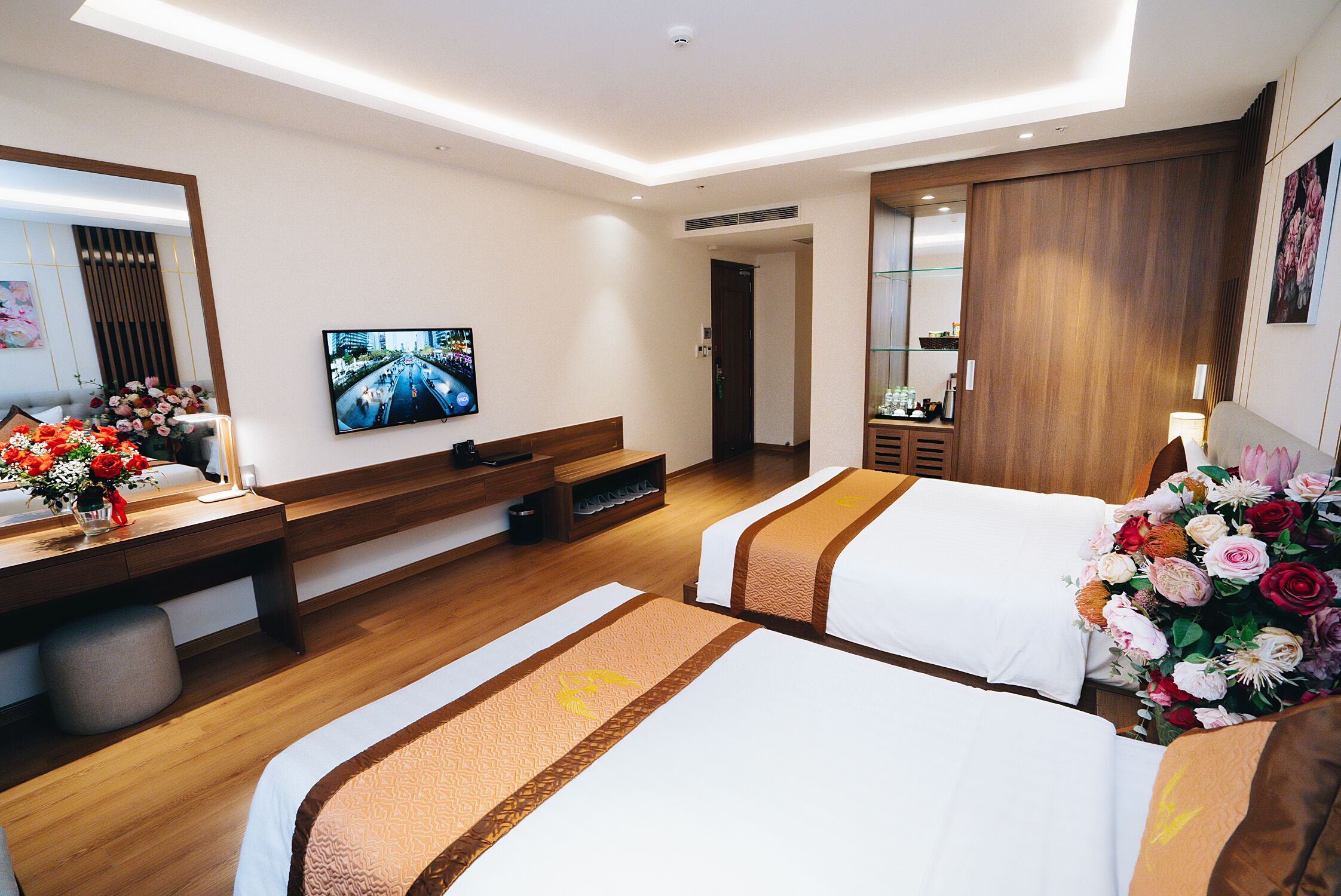 A hotel room with two beds, a TV, a desk with a vase of flowers, and a wooden wardrobe.