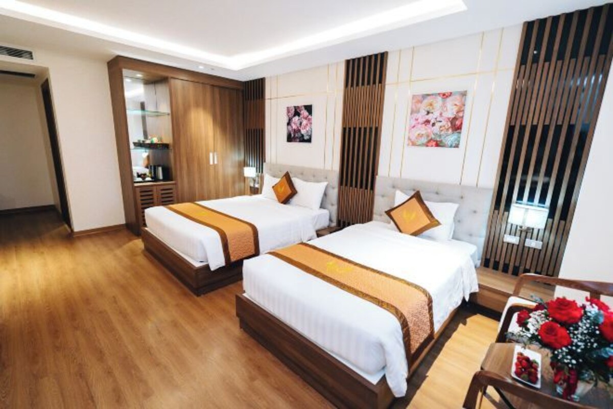 Habitación de hotel con dos camas, suelo de madera, un escritorio con silla y un florero con flores.
