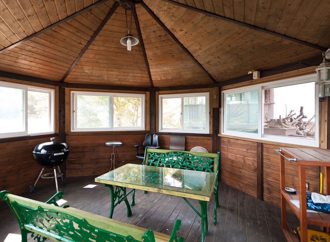 Uma área externa coberta com teto de madeira, mesa de vidro, bancos verdes, churrasqueira e vista para o exterior.