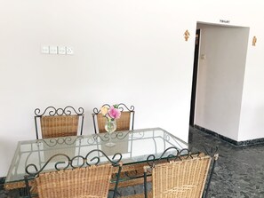 A dining area with a glass table, wicker chairs, and a vase with flowers. There is a white wall with a power outlet and a door labeled "TOILET".