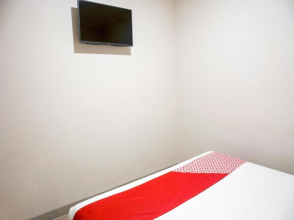 Una habitación con una cama, un pequeño televisor montado en la pared y una colcha de edredón con motivos rojos y blancos.