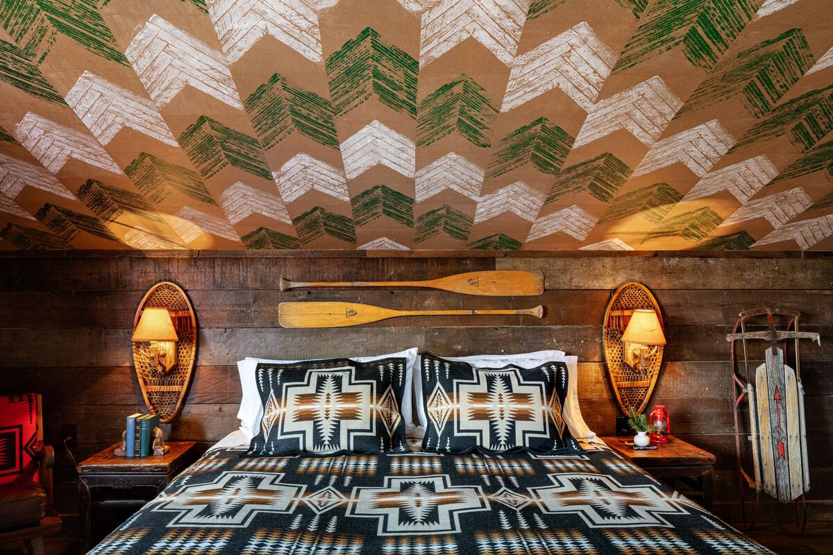 A room with a patterned ceiling, wooden walls, a bed with patterned bedding, two wall-mounted lamps, and a wooden bat on the wall.