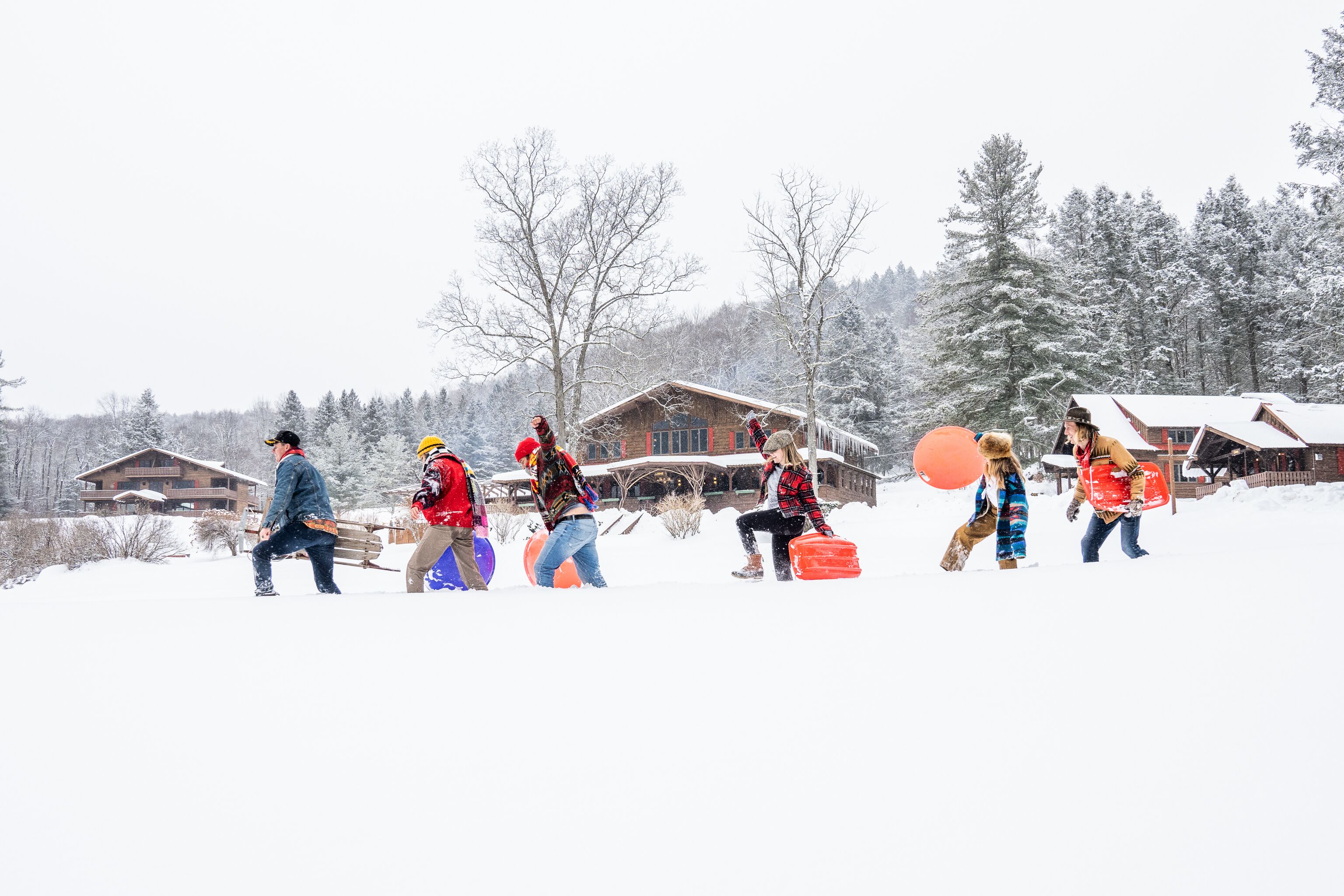 Un groupe de personnes, vêtues pour l’hiver, transporte des planches à neige et une luge, et marche sur un sentier enneigé. On aperçoit un chalet en bois au loin.