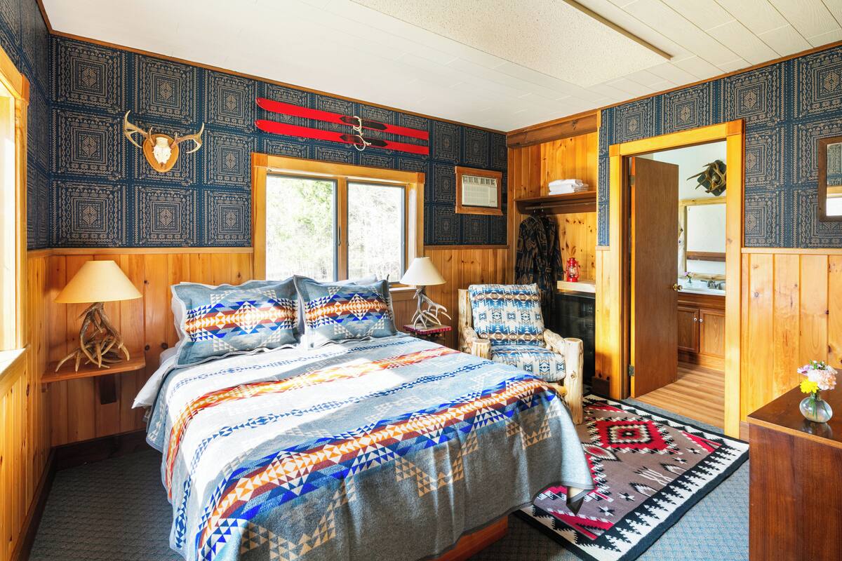 A wooden-themed bedroom with a bed, two chairs, a nightstand, and a window.