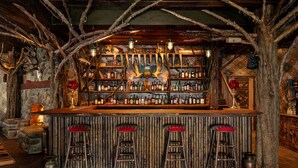 A rustic bar area with wooden stools, a wooden bar counter, and a backdrop of antlers and a welcome sign.