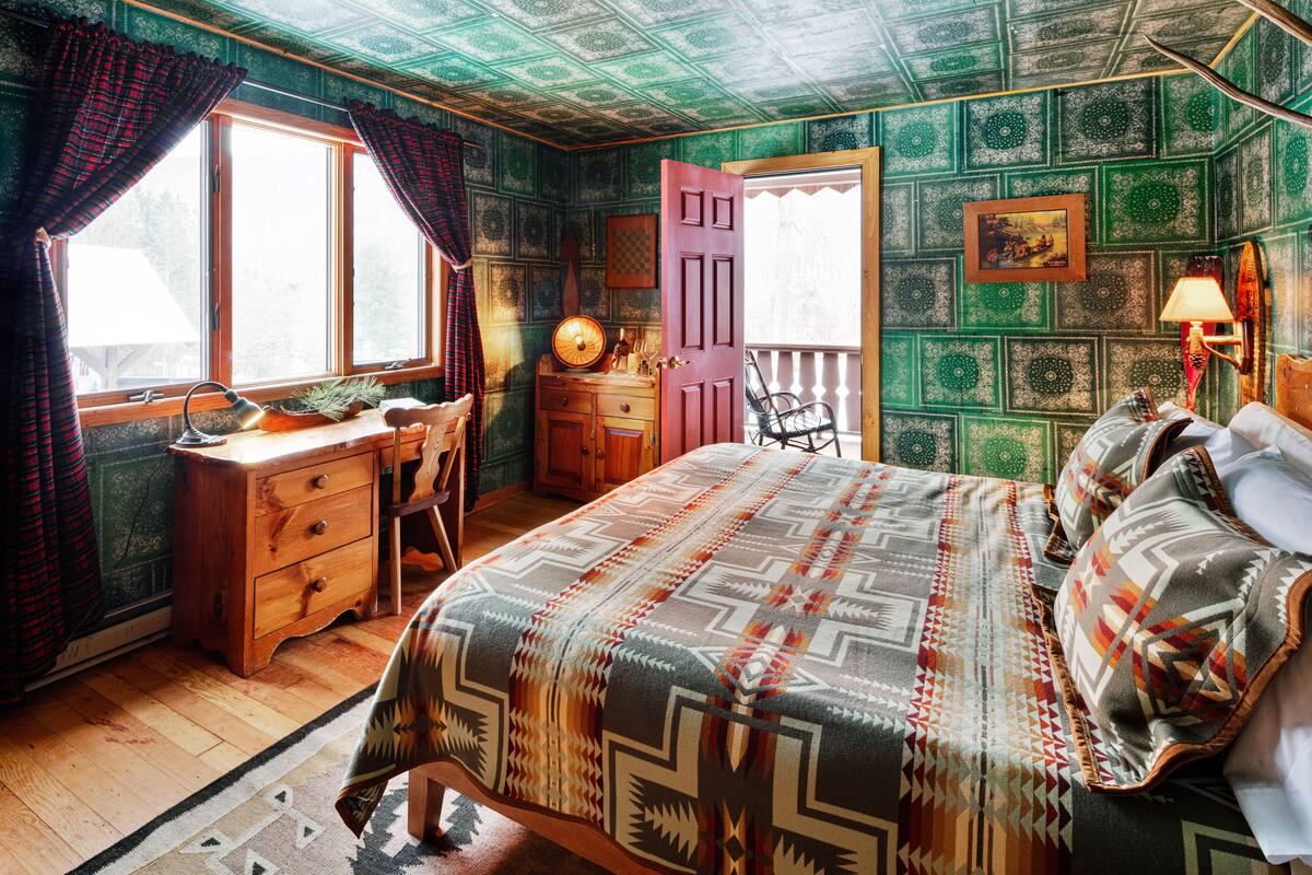 A cozy room with a bed, a desk, and a chair. There is a window with curtains, a lamp, and a door leading outside.