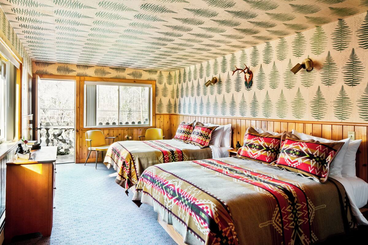 A room with two beds, a desk, and a chair. The walls have a leaf pattern, and there is a window with a view of trees.