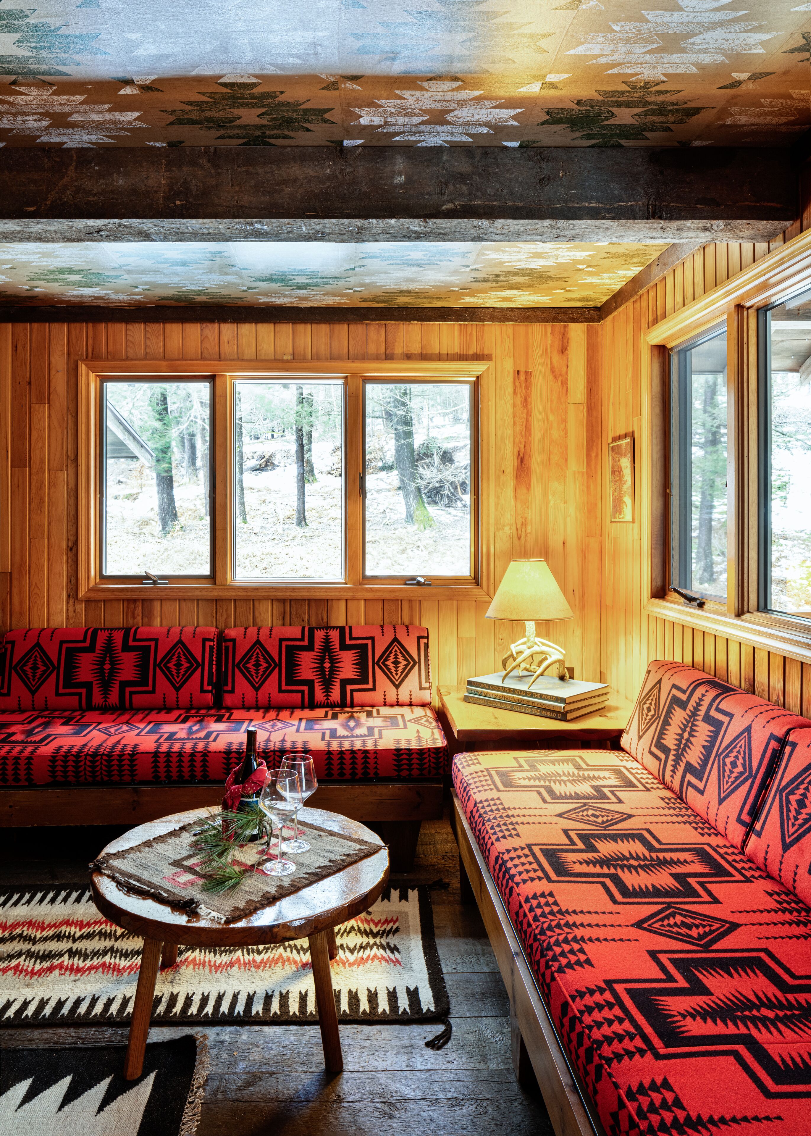 A cozy cabin interior with a wooden interior, a sofa, a table with a bottle and glasses, and a window with a view of trees.