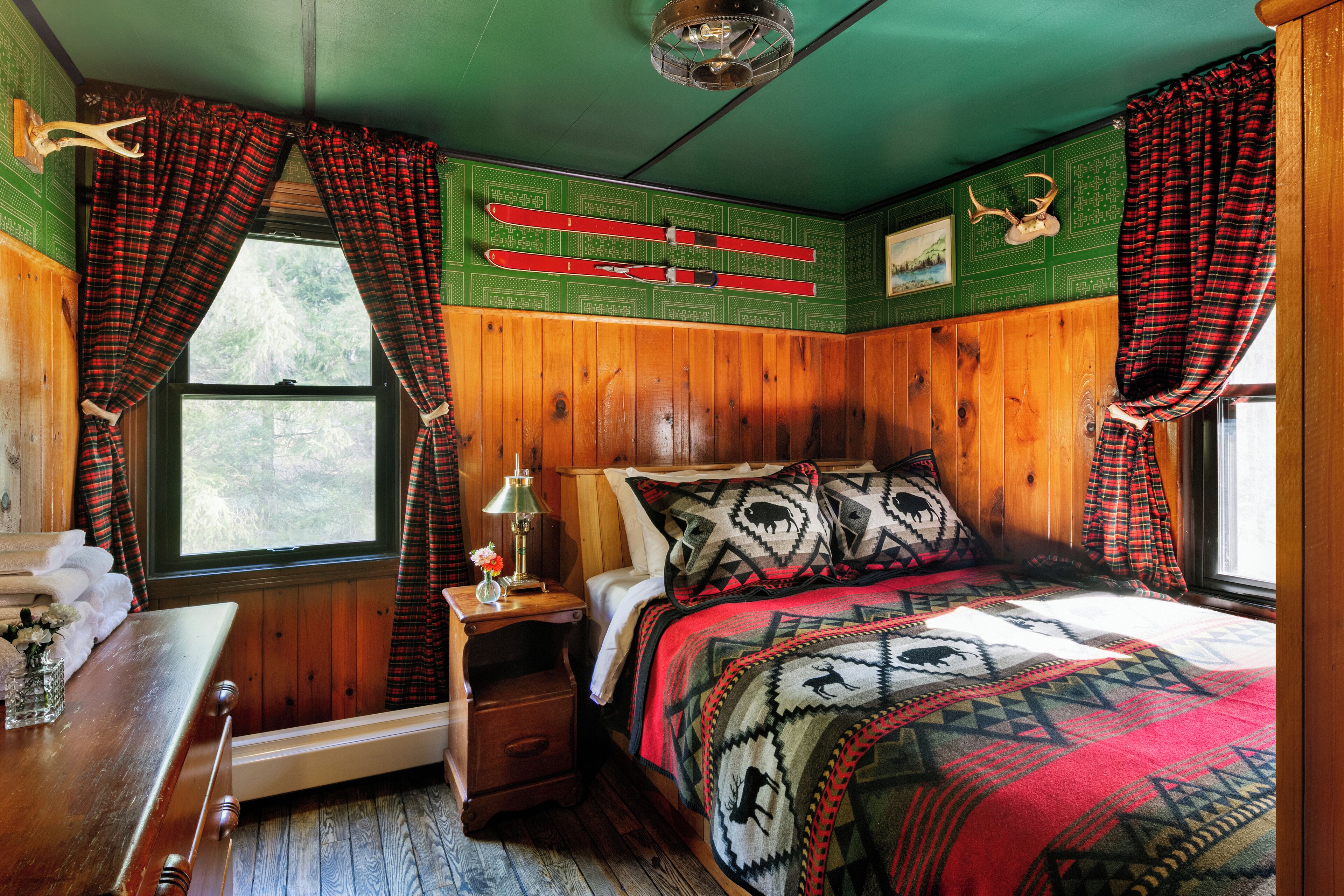 Une chambre confortable dans un chalet, avec des murs en bois, un lit recouvert d’une literie à motifs, une table de chevet avec une lampe et une fenêtre avec des rideaux.