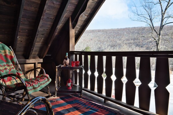 A wooden deck with a rocking chair and a table with a lantern. A view of a forested mountain is visible through a railing. - Urban Cowboy Lodge (Big Indian)