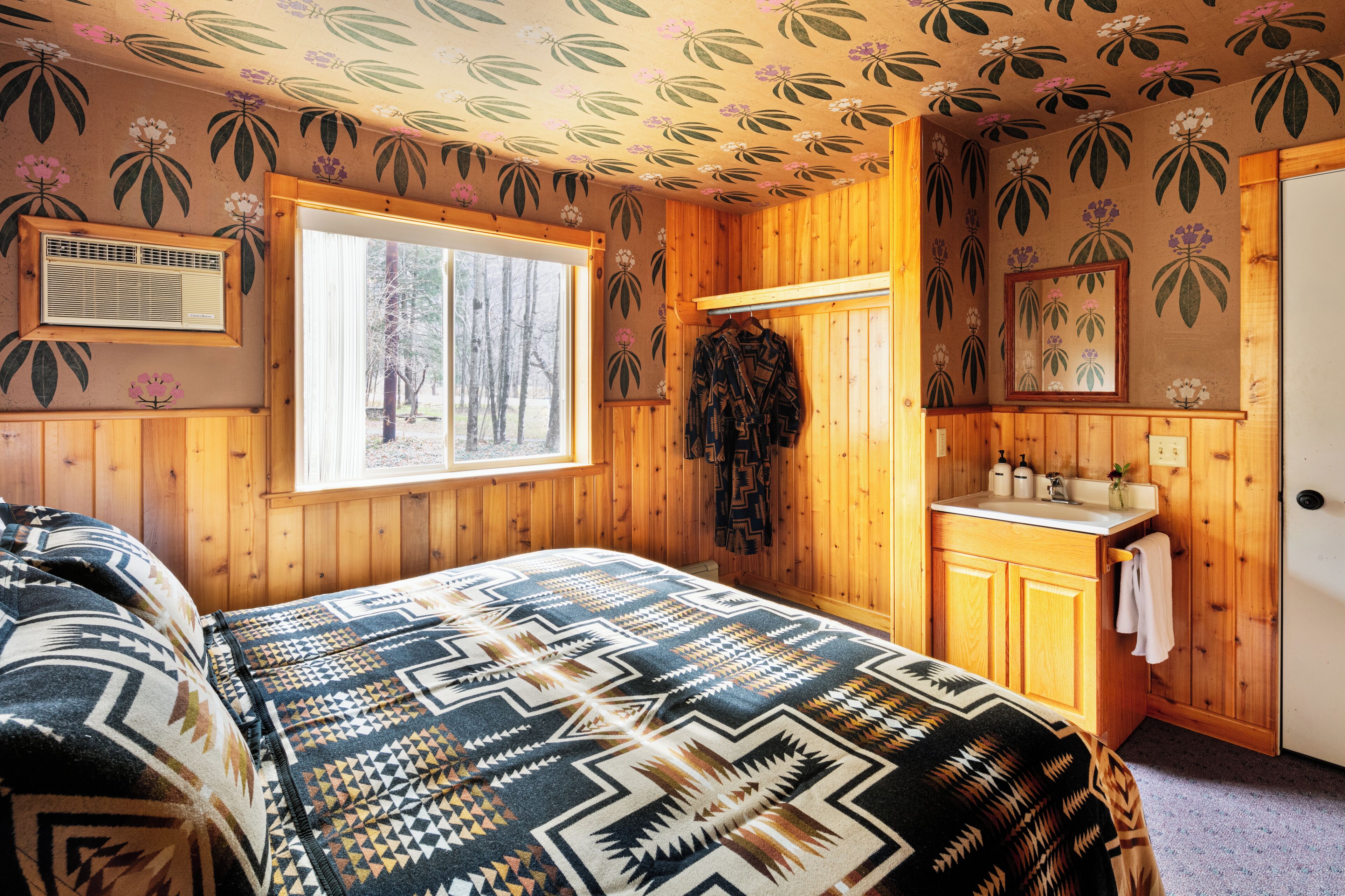 A cozy room with a bed, a window with a view of trees, a wooden wall with floral wallpaper, and a small bathroom with a sink and mirror.