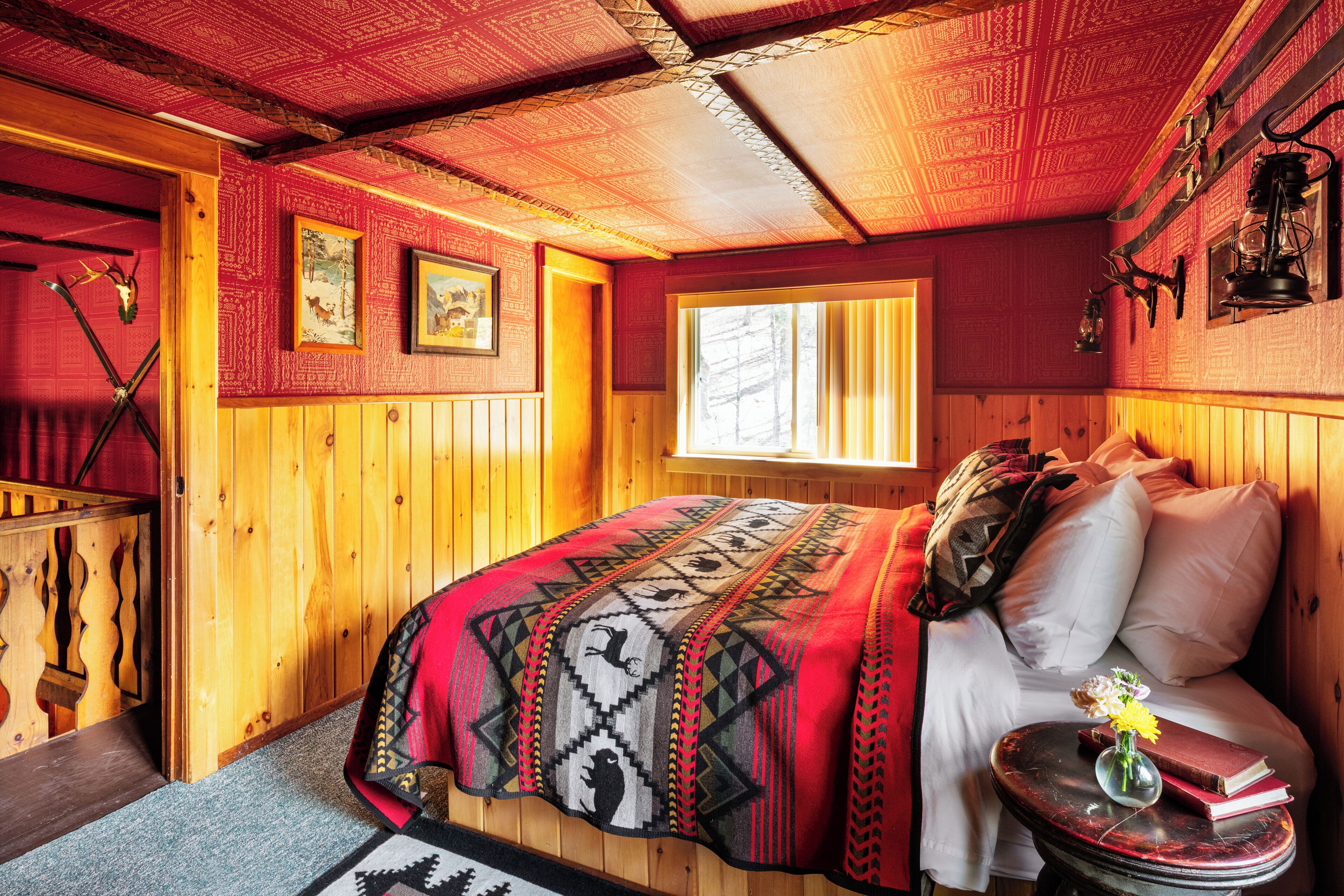 Una habitación acogedora con paredes de madera, una cama con un edredón estampado, una ventana y una mesita con un jarrón y un libro.
