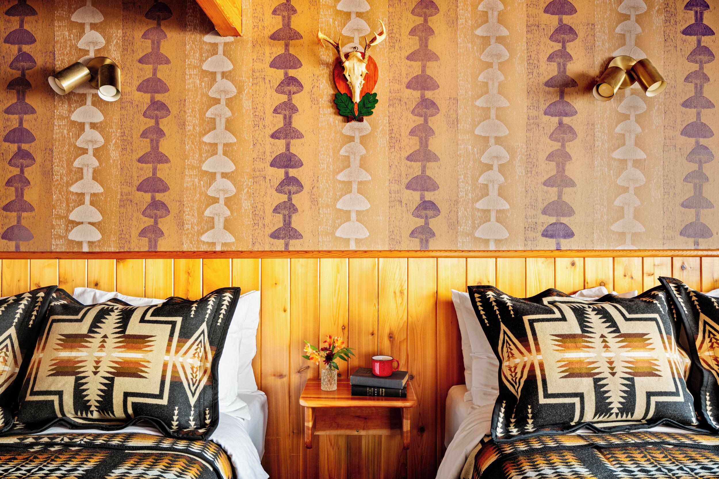 A room with two beds, a wooden headboard, a nightstand with a red mug and books, and a wall with a patterned design.
